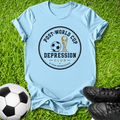 A light blue short-sleeve t-shirt laid flat on green grass, featuring a circular graphic that reads 'POST-WORLD CUP DEPRESSION CLUB' with a soccer ball and trophy illustration. A black-and-white soccer ball appears at the lower left, and black cleats are at the lower right.
