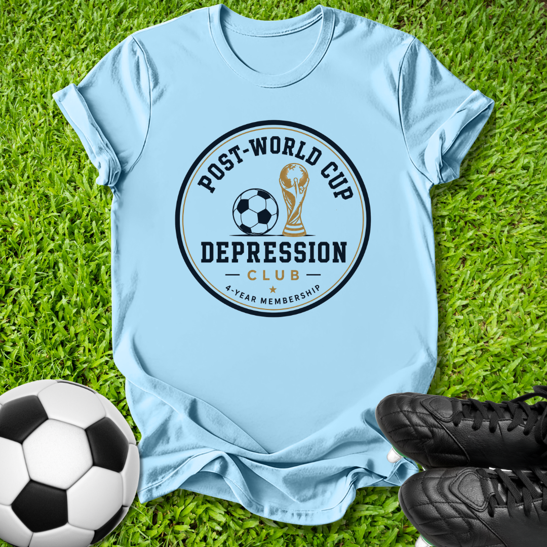 A light blue short-sleeve t-shirt laid flat on green grass, featuring a circular graphic that reads 'POST-WORLD CUP DEPRESSION CLUB' with a soccer ball and trophy illustration. A black-and-white soccer ball appears at the lower left, and black cleats are at the lower right.