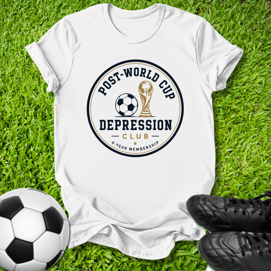 A white short-sleeve T-shirt laid flat on green grass, featuring a circular graphic that reads 'POST-WORLD CUP DEPRESSION CLUB' with a soccer ball, trophy, and '4 YEAR MEMBERSHIP' text. A black-and-white soccer ball appears at the lower left, and black cleats are at the lower right.