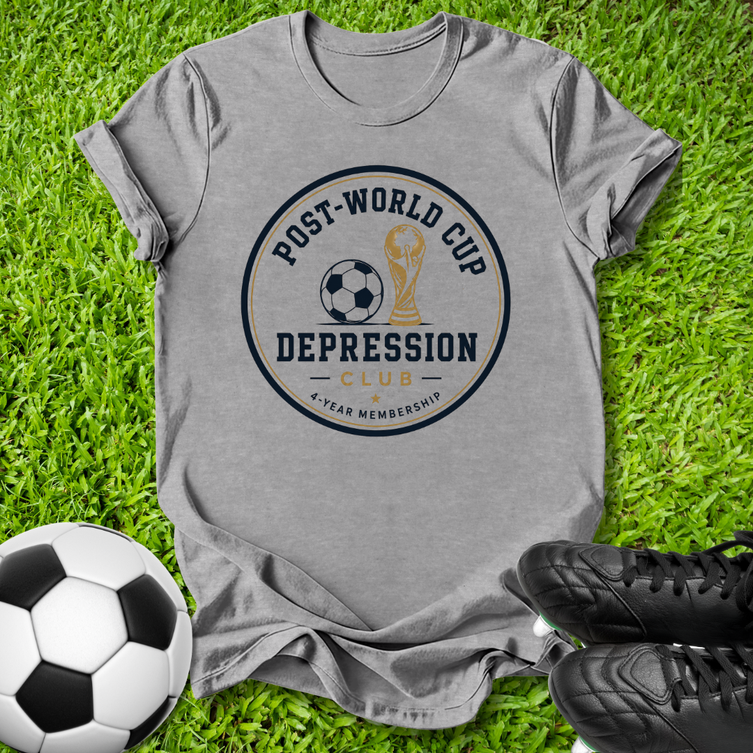 A heather gray graphic t-shirt laid flat on green grass, featuring a circular print that reads 'POST-WORLD CUP DEPRESSION CLUB' with a soccer ball and trophy illustration. A soccer ball and black cleats are placed around it as props.