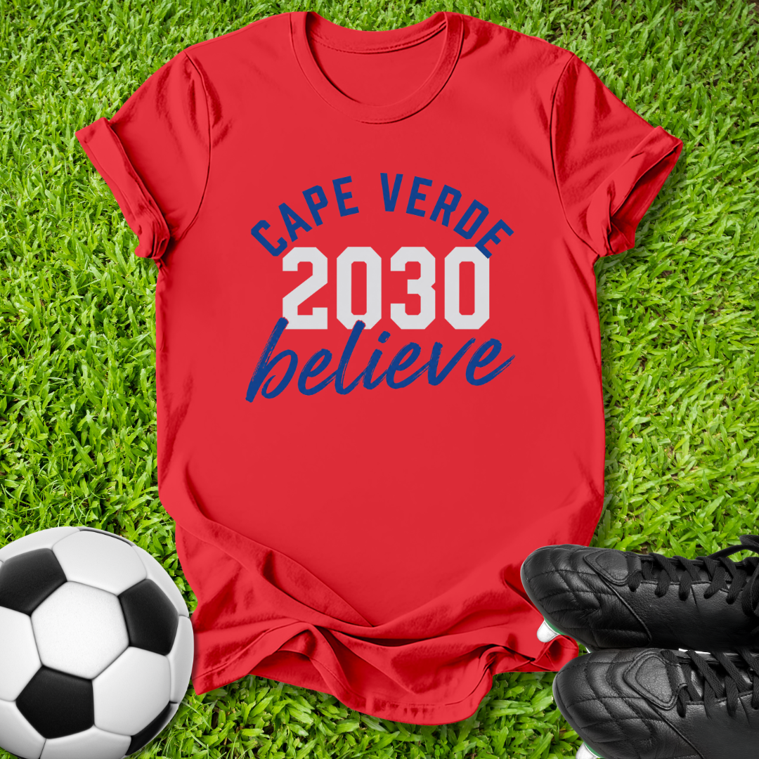 A red short-sleeve graphic T-shirt laid flat on green grass, with a soccer ball at the lower left and black cleats at the lower right. The shirt reads 'CAPE VERDE 2030 believe' in blue and white text.