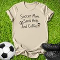 A beige graphic t-shirt laid flat on green grass, featuring the text 'Soccer Mom. Send Help.