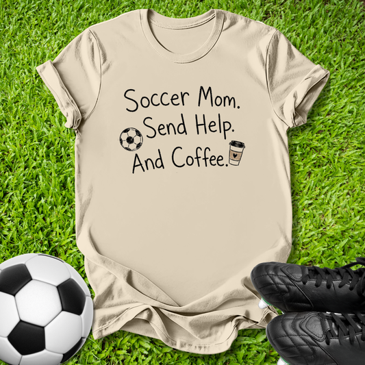 A beige graphic t-shirt laid flat on green grass, featuring the text 'Soccer Mom. Send Help.