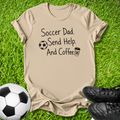 A beige graphic t-shirt laid flat on green grass, featuring the text 'Soccer Dad. Send Help.