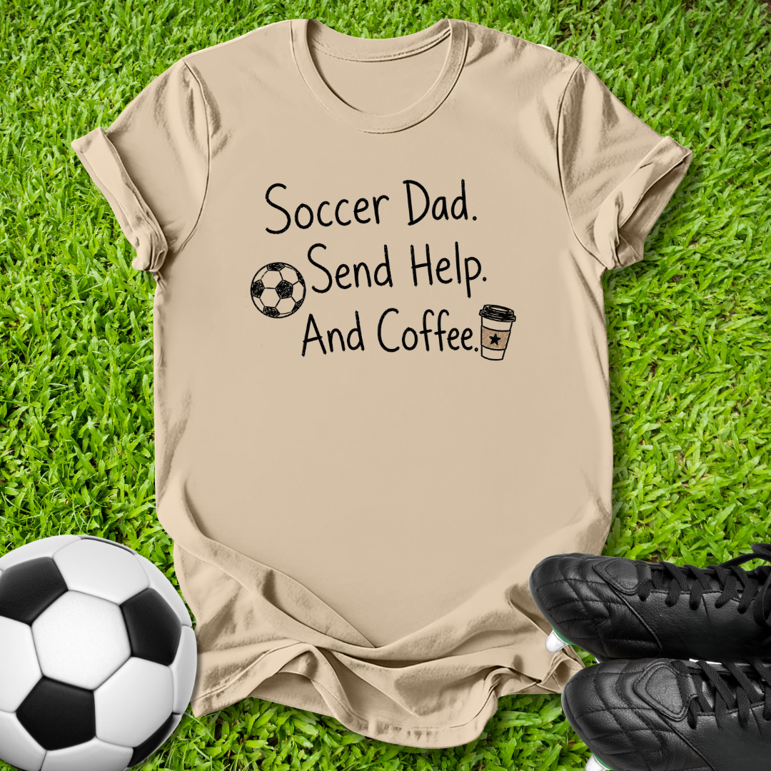 A beige graphic t-shirt laid flat on green grass, featuring the text 'Soccer Dad. Send Help.