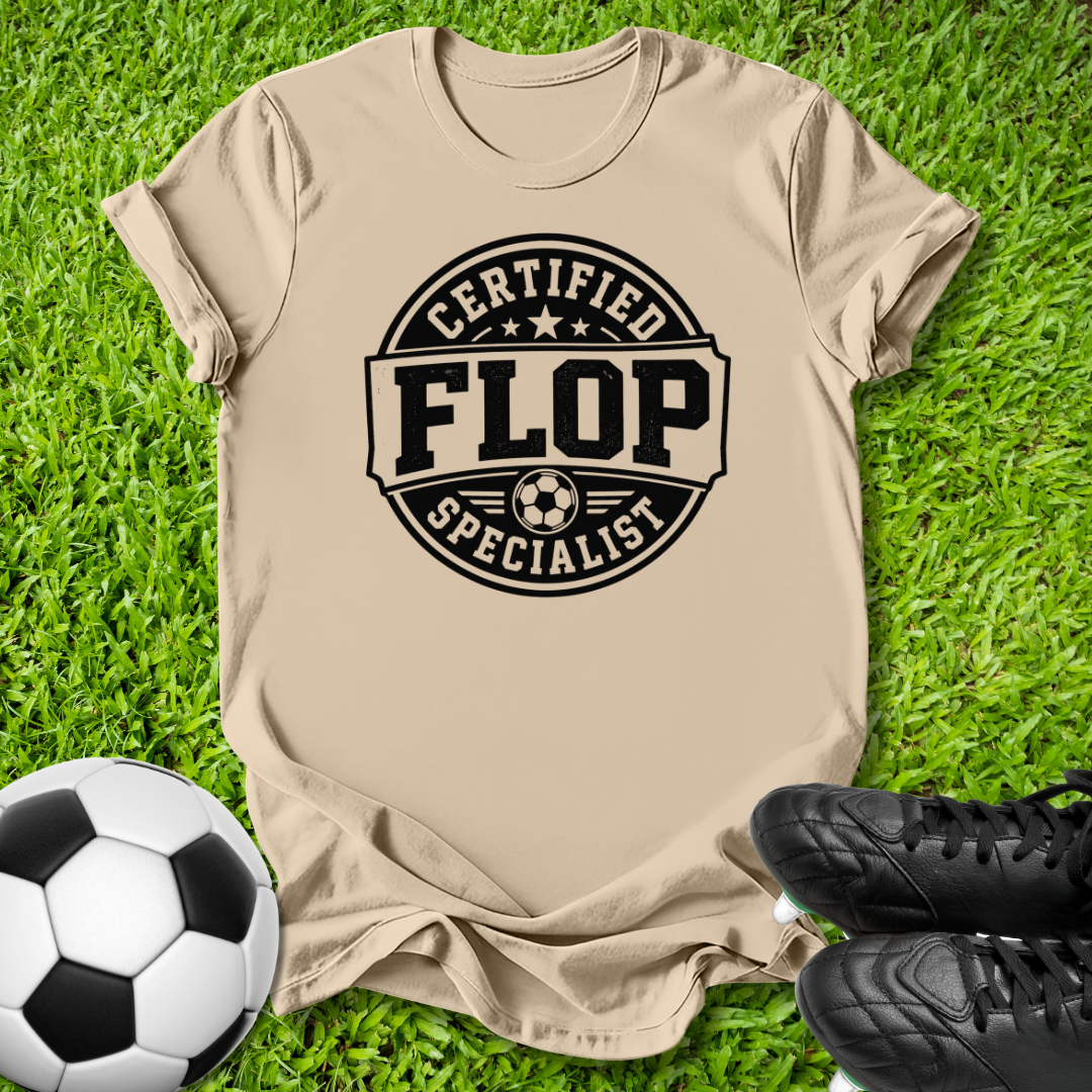 A beige graphic t-shirt laid flat on green grass, featuring a black circular stamp design that reads 'CERTIFIED FLOP SPECIALIST' with a soccer ball icon. A black-and-white soccer ball is on the left, and black cleats are on the right.