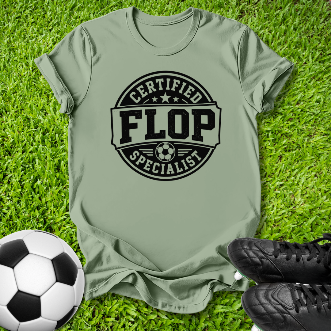 An olive green t-shirt with a black graphic reading 'CERTIFIED FLOP SPECIALIST' and a soccer ball icon, laid flat on green grass with a soccer ball and black cleats as props.