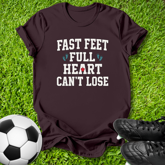 A dark brown short-sleeve t-shirt with the text 'FAST FEET FULL HEART CAN'T LOSE' printed in white, styled on green grass with a soccer ball and black cleats.