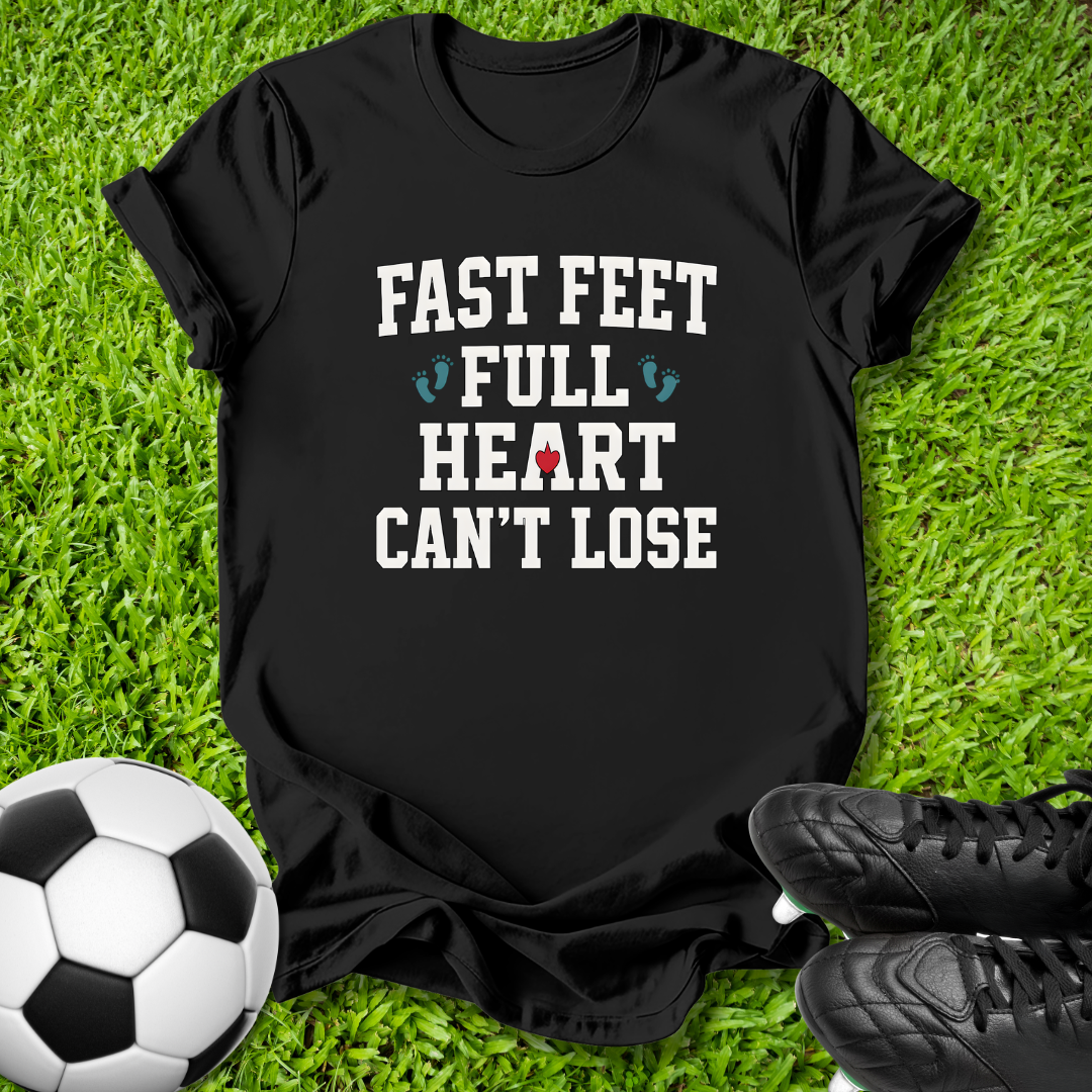 A black short-sleeve graphic T-shirt laid flat on green grass, with a soccer ball at the lower left and black cleats at the lower right. The shirt reads “FAST FEET FULL HEART CAN’T LOSE” in white text with small blue and red graphic accents.