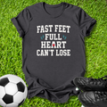 A black graphic t-shirt laid flat on green grass, featuring the text 'FAST FEET FULL HEART CAN'T LOSE' in white with small blue and red accents. A soccer ball is on the left and black cleats are on the right.