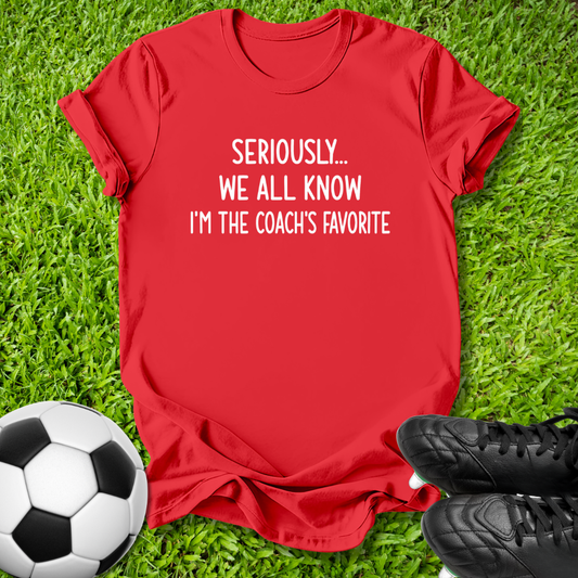 A red short-sleeve graphic T-shirt laid flat on green grass, with a soccer ball on the left and black cleats on the right. The shirt has white text reading 'SERIOUSLY.