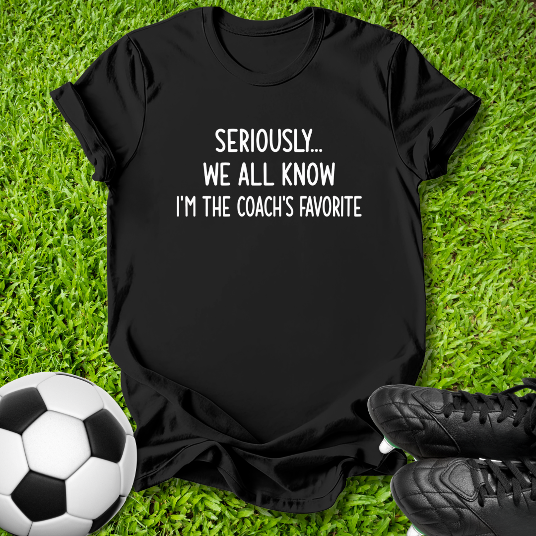 A black short-sleeve graphic T-shirt laid flat on green grass, with a soccer ball on the left and black cleats on the right. The shirt has white text reading 'SERIOUSLY.