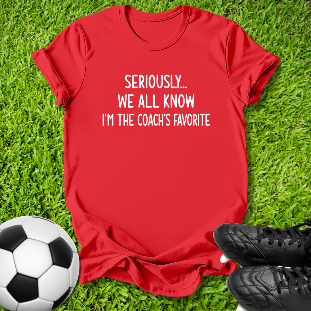 A red short-sleeve graphic T-shirt laid flat on green grass, with a soccer ball on the left and black cleats on the right. The shirt has white text reading 'SERIOUSLY.
