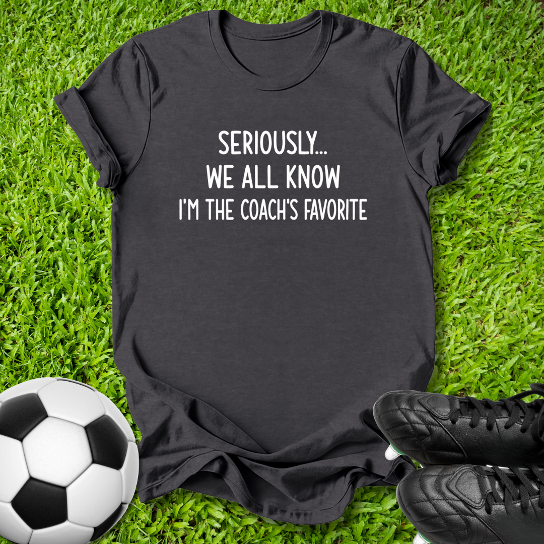 A black short-sleeve graphic t-shirt laid flat on green grass, with a soccer ball on the left and black cleats on the right. The shirt features the white printed text 'SERIOUSLY.