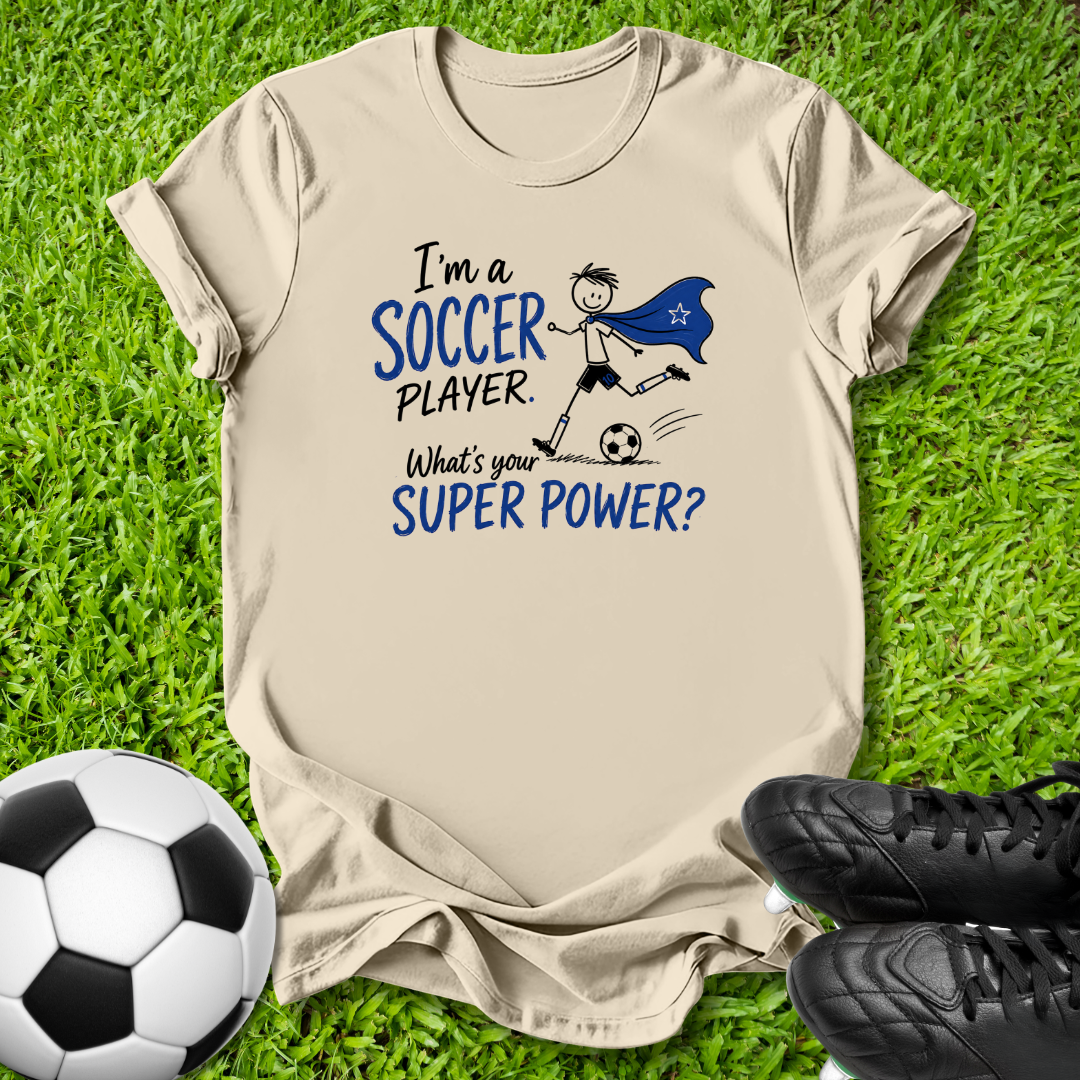 A beige graphic t-shirt laid flat on green grass, featuring a blue and black cartoon soccer player design with the text 'I'm a SOCCER PLAYER. What's your SUPER POWER?'.