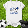 A white short-sleeve graphic T-shirt laid flat on green grass, with a soccer-themed print reading 'I'm a SOCCER PLAYER. What's your SUPER POWER?' in blue and black text, plus a simple soccer player illustration.