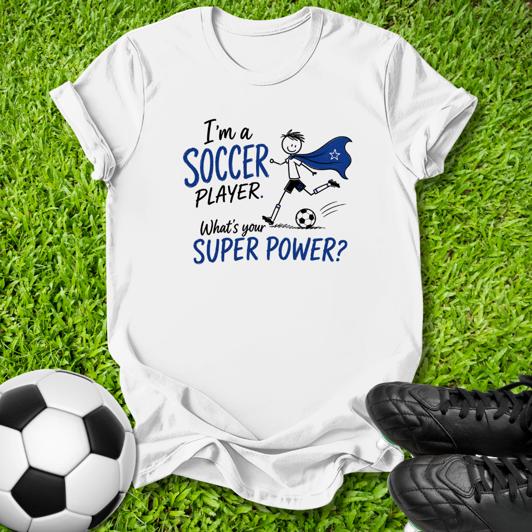A white short-sleeve graphic T-shirt laid flat on green grass, with a soccer-themed print reading 'I'm a SOCCER PLAYER. What's your SUPER POWER?' in blue and black text, plus a simple soccer player illustration.