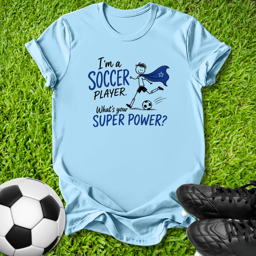 A light blue graphic t-shirt laid flat on green grass, featuring a black and blue cartoon illustration of a soccer player in a superhero cape with the text 'I'm a SOCCER PLAYER. What's your SUPER POWER?'.
