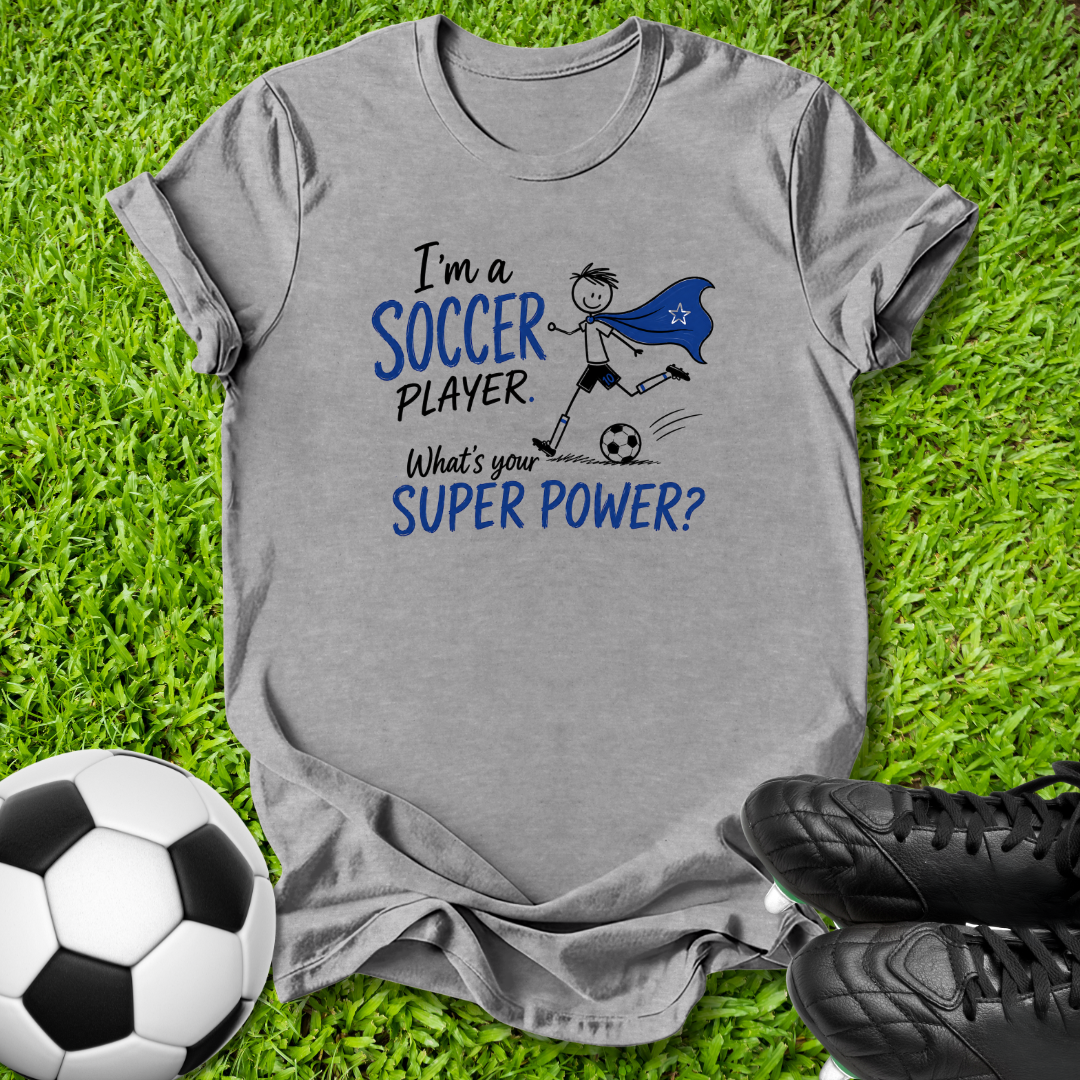 A heather gray graphic t-shirt laid flat on green grass, featuring a blue and black print that reads 'I'm a SOCCER PLAYER. What's your SUPER POWER?' with a cartoon soccer player in a cape.