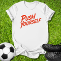 A white short-sleeve t-shirt with the phrase 'PUSH YOURSELF' printed in red across the chest, laid flat on green grass with a soccer ball and black cleats as props.