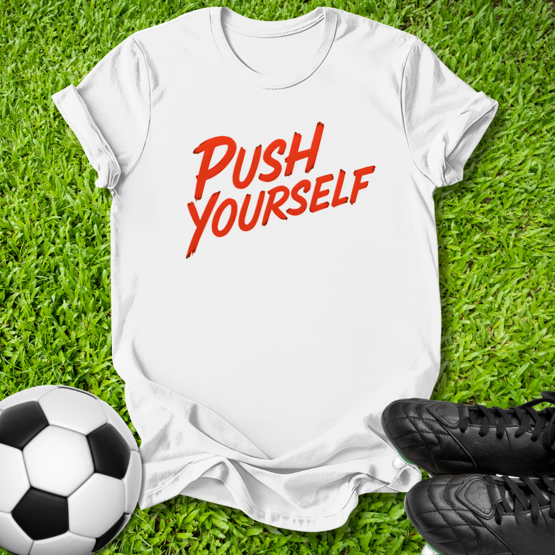 A white short-sleeve t-shirt with the phrase 'PUSH YOURSELF' printed in red across the chest, laid flat on green grass with a soccer ball and black cleats as props.