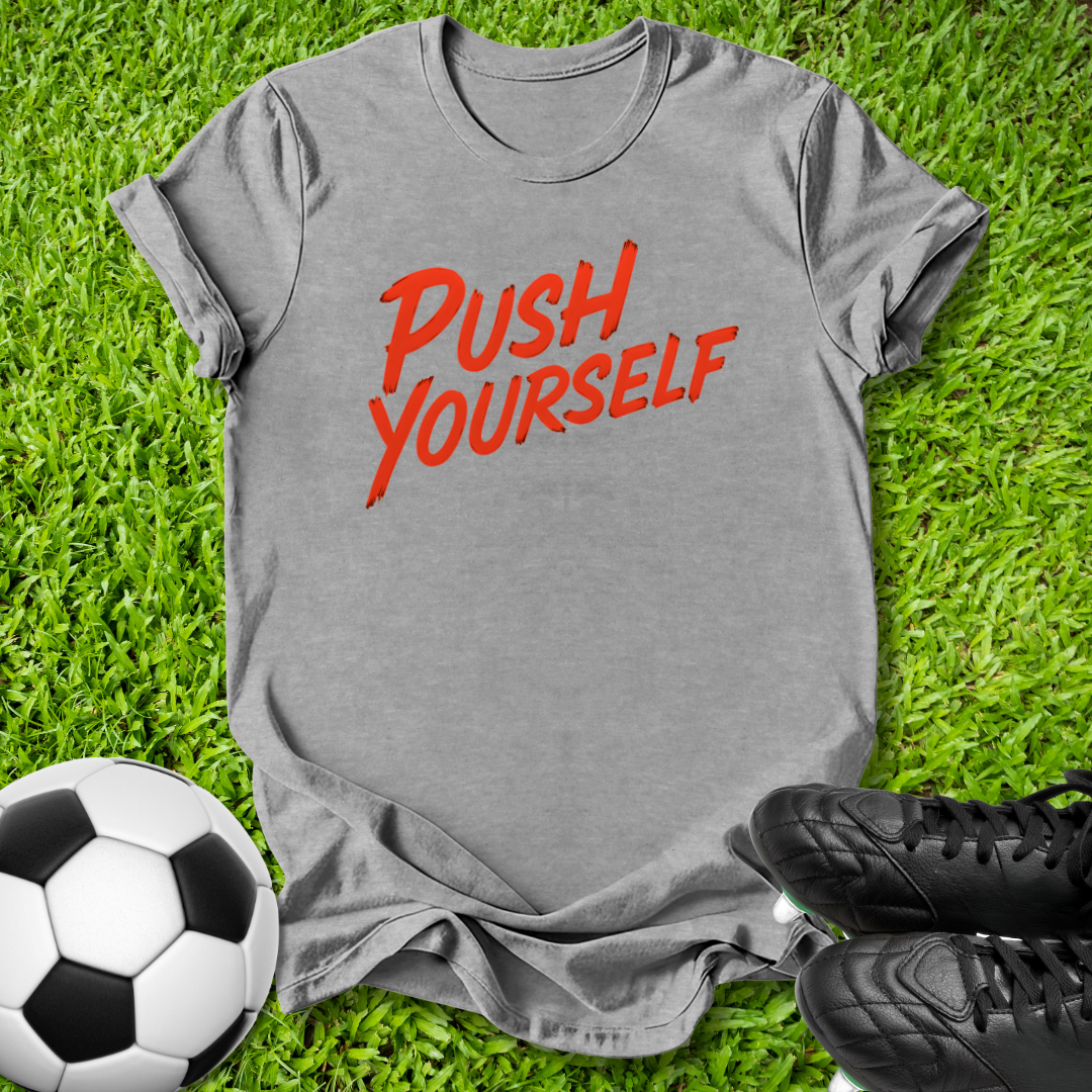 A heather gray short-sleeve t-shirt with the phrase 'PUSH YOURSELF' printed in red across the chest, laid flat on green grass with a soccer ball and black cleats as props.