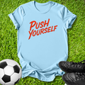 A light blue t-shirt with the words 'PUSH YOURSELF' printed in red across the chest, laid flat on green grass with a soccer ball and black cleats as props.