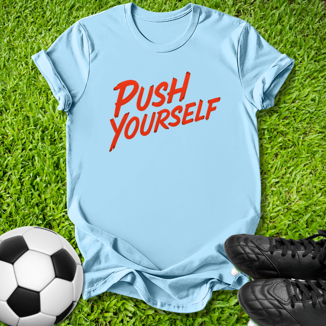 A light blue t-shirt with the words 'PUSH YOURSELF' printed in red across the chest, laid flat on green grass with a soccer ball and black cleats as props.