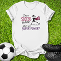 A white graphic t-shirt laid flat on green grass, featuring a soccer-themed design with the text 'I'm a SOCCER PLAYER. What's your SUPER POWER?' and a simple illustration of a soccer player with a cape.