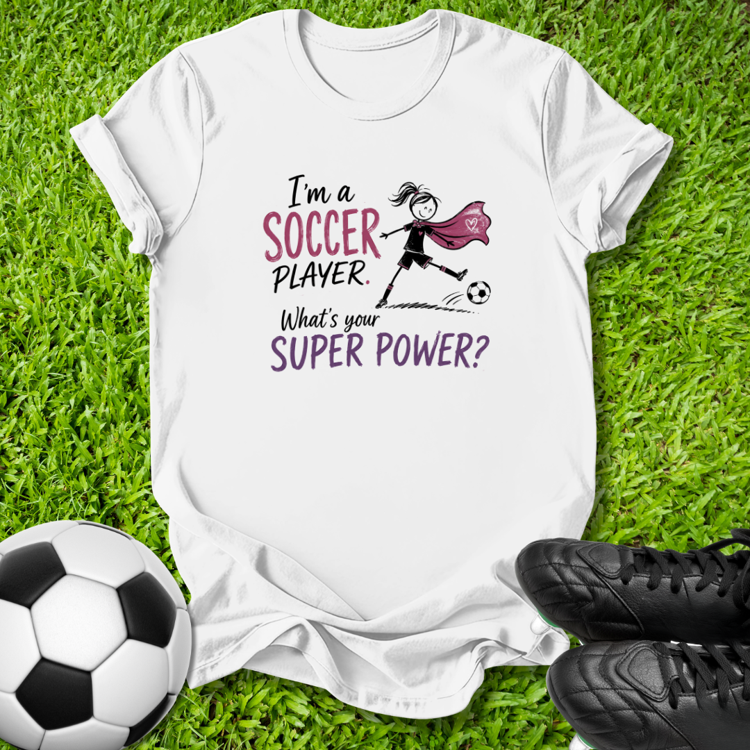 A white graphic t-shirt laid flat on green grass, featuring a soccer-themed design with the text 'I'm a SOCCER PLAYER. What's your SUPER POWER?' and a simple illustration of a soccer player with a cape.