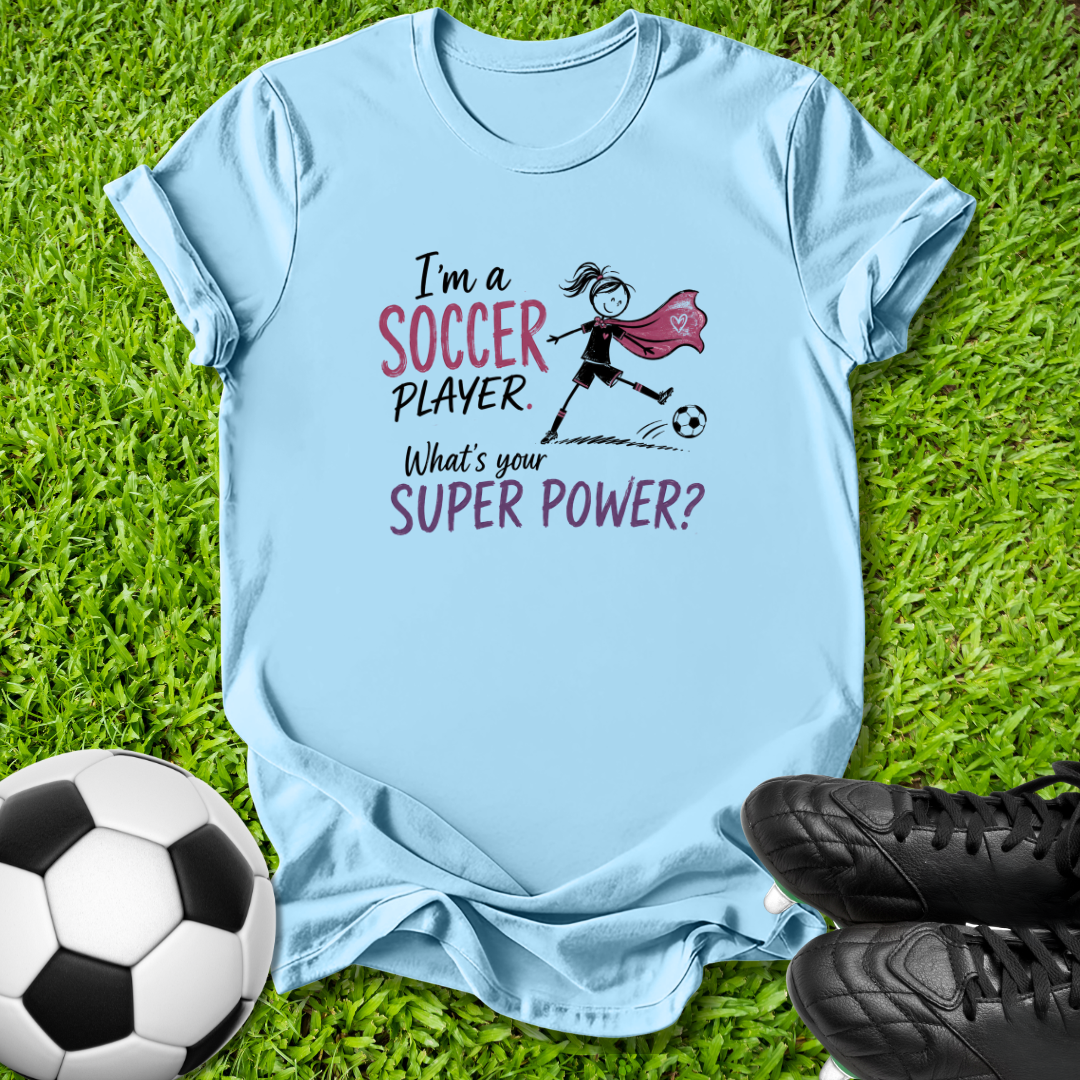 A light blue graphic t-shirt laid flat on green grass, featuring a soccer-themed design with the text 'I'm a SOCCER PLAYER. What's your SUPER POWER?' and an illustration of a girl in a black dress with a pink cape kicking a soccer ball.