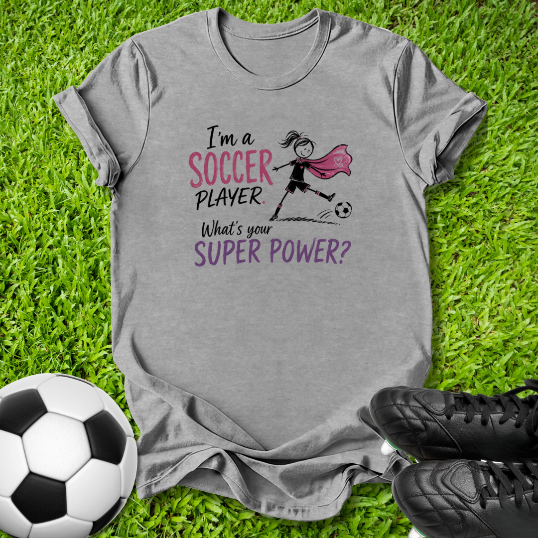 A heather gray graphic t-shirt laid flat on green grass, featuring a soccer-themed design with the text 'I'm a SOCCER PLAYER What's your SUPER POWER?' and a simple illustration of a soccer player with a pink cape. A soccer ball and black cleats are placed around the shirt as props.