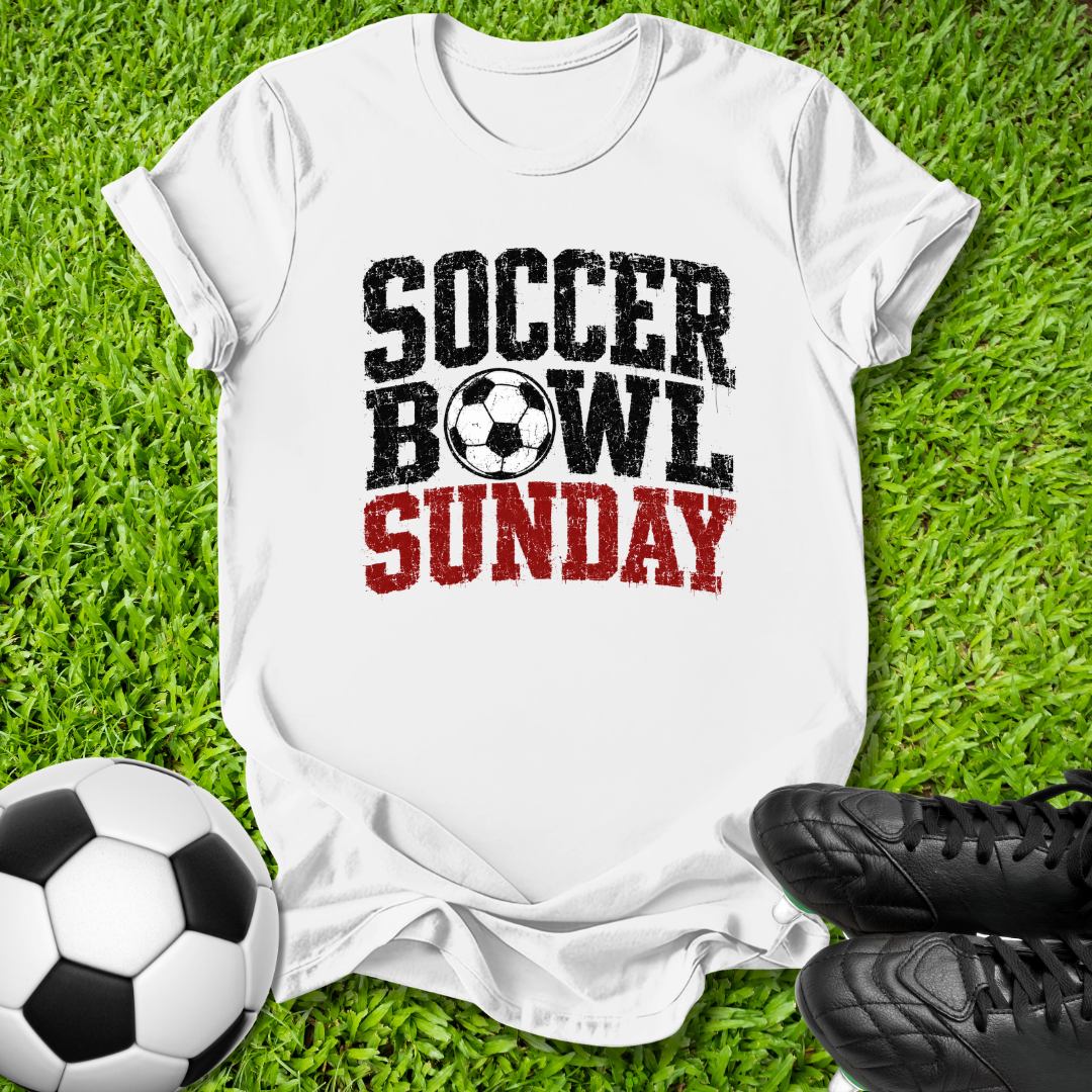A white short-sleeve graphic T-shirt laid flat on green grass, with a soccer ball at the lower left and black cleats at the lower right. The shirt reads 'SOCCER BOWL SUNDAY' in distressed black and red lettering, with a soccer ball icon between 'BOWL' and 'SUNDAY'.