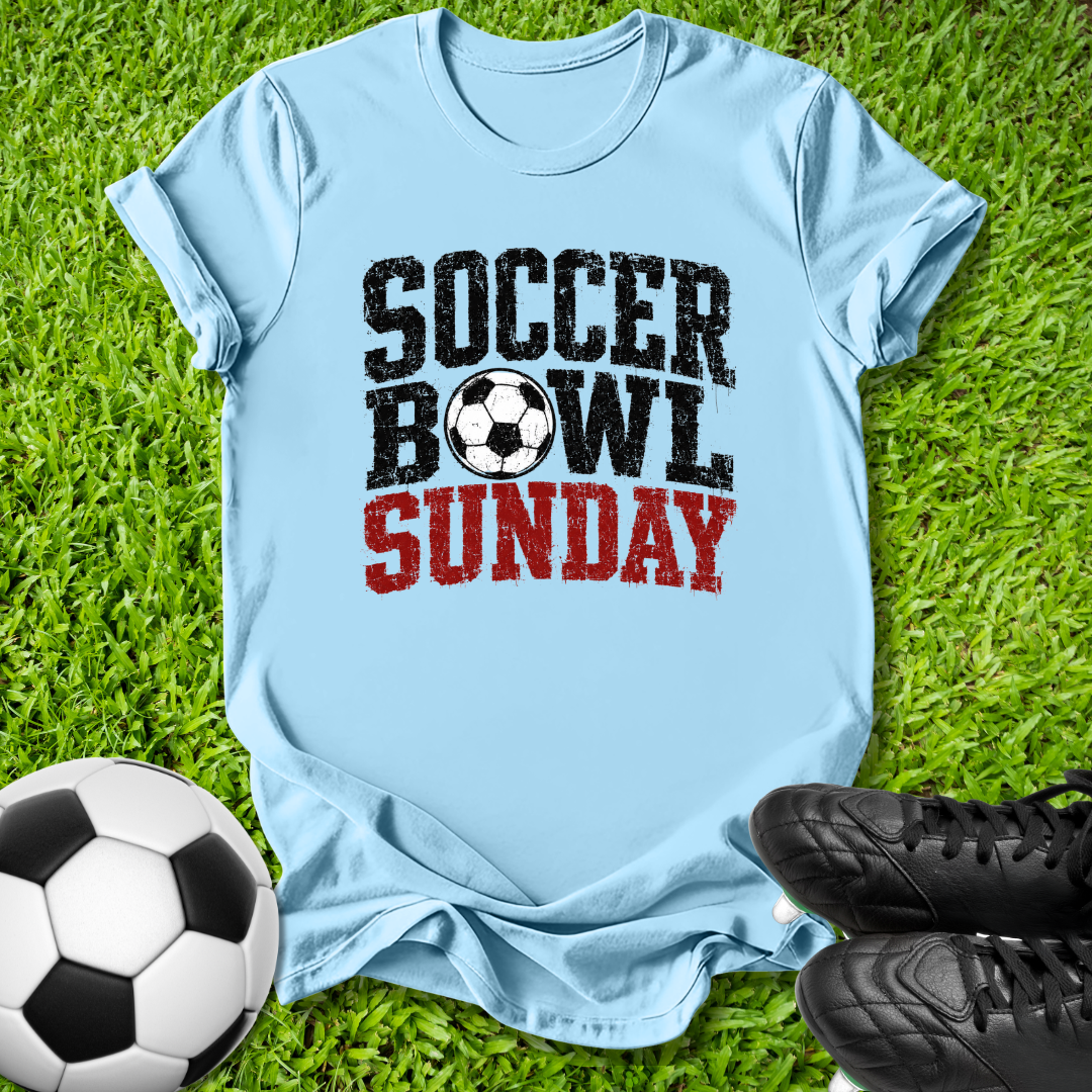 A light blue graphic t-shirt laid flat on green grass, featuring the text 'SOCCER BOWL SUNDAY' in black and red with a soccer ball graphic. A soccer ball and black cleats are placed around it as props.