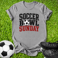 A heather gray graphic t-shirt laid flat on green grass, featuring the text 'SOCCER BOWL SUNDAY' in distressed black and red lettering with a soccer ball graphic. A soccer ball and black cleats are styled around the shirt.