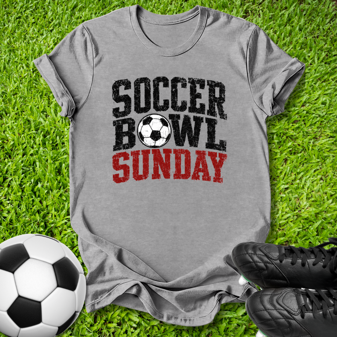 A heather gray graphic t-shirt laid flat on green grass, featuring the text 'SOCCER BOWL SUNDAY' in distressed black and red lettering with a soccer ball graphic. A soccer ball and black cleats are styled around the shirt.