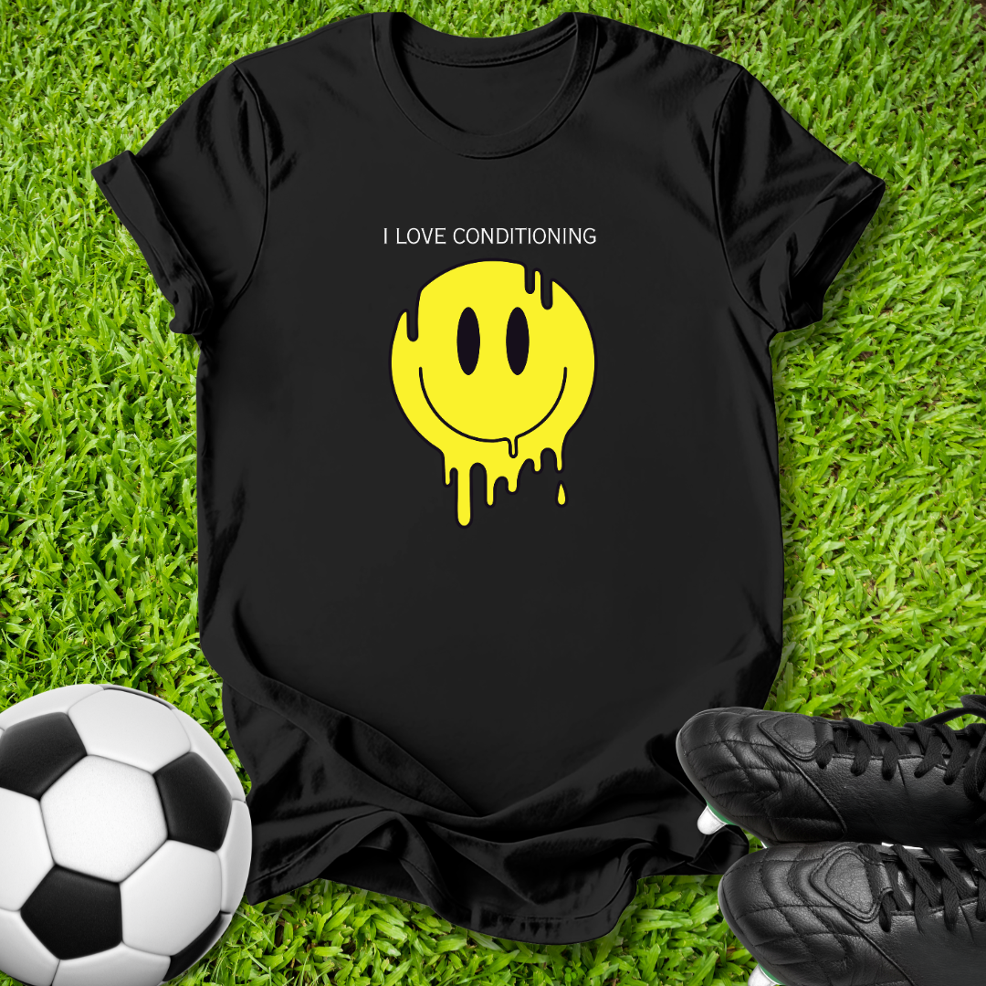A black short-sleeve T-shirt laid flat on green grass, featuring a yellow dripping smiley face graphic with the text 'I LOVE CONDITIONING' above it. A soccer ball is visible at the lower left and black cleats at the lower right.