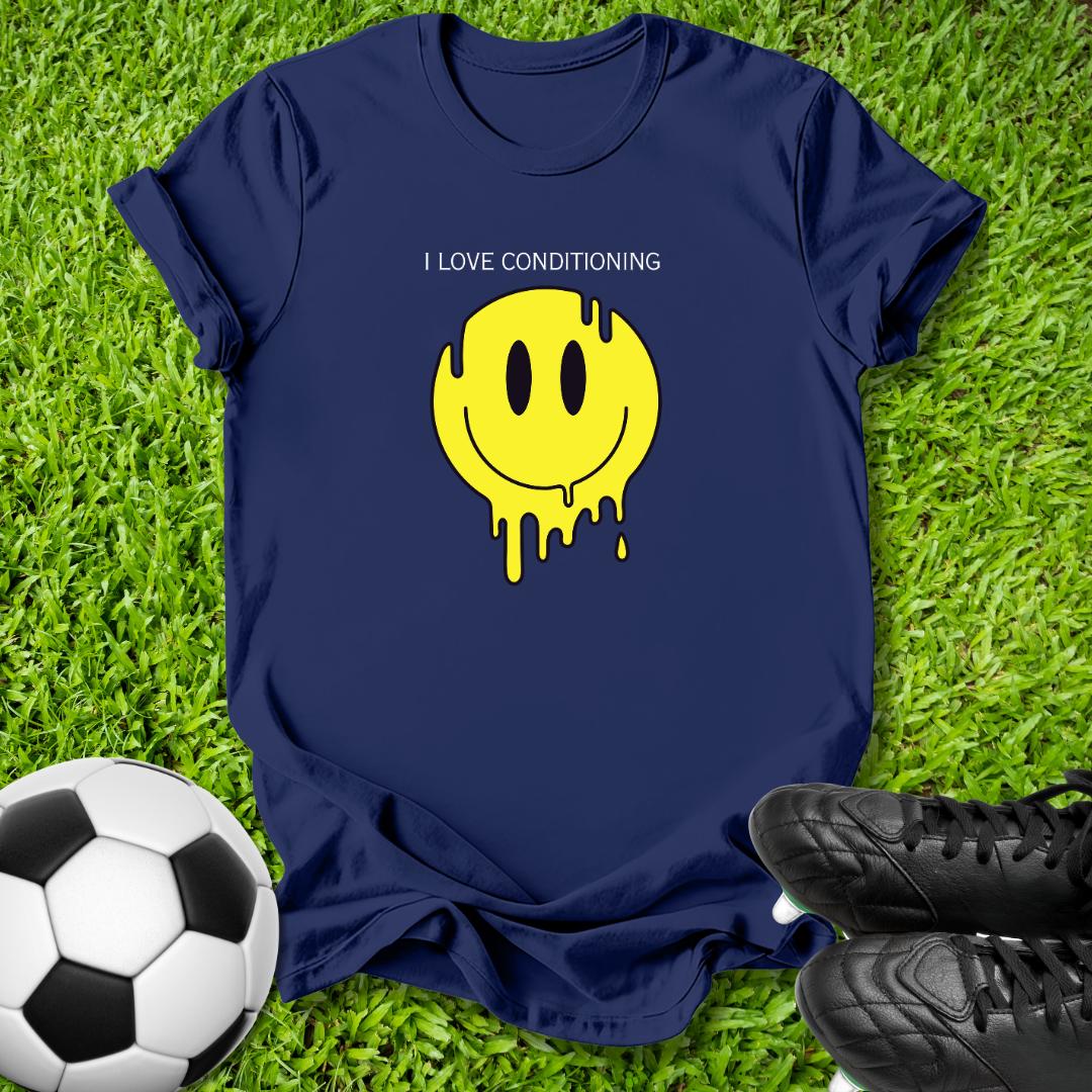 A navy blue t-shirt laid flat on green grass, featuring a yellow dripping smiley face graphic with the text 'I LOVE CONDITIONING' above it. A soccer ball is visible on the left and black cleats are visible on the right, creating a sports-themed flat-lay composition.