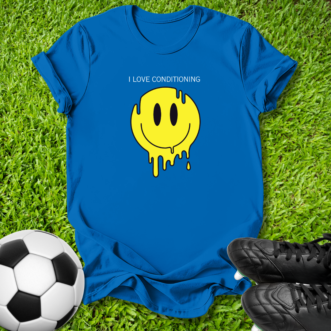 A royal blue short-sleeve t-shirt laid flat on green grass, featuring a yellow dripping smiley face graphic with the text 'I LOVE CONDITIONING' above it. A soccer ball is visible on the left and black cleats are visible on the right, creating a casual outdoor flat-lay scene.