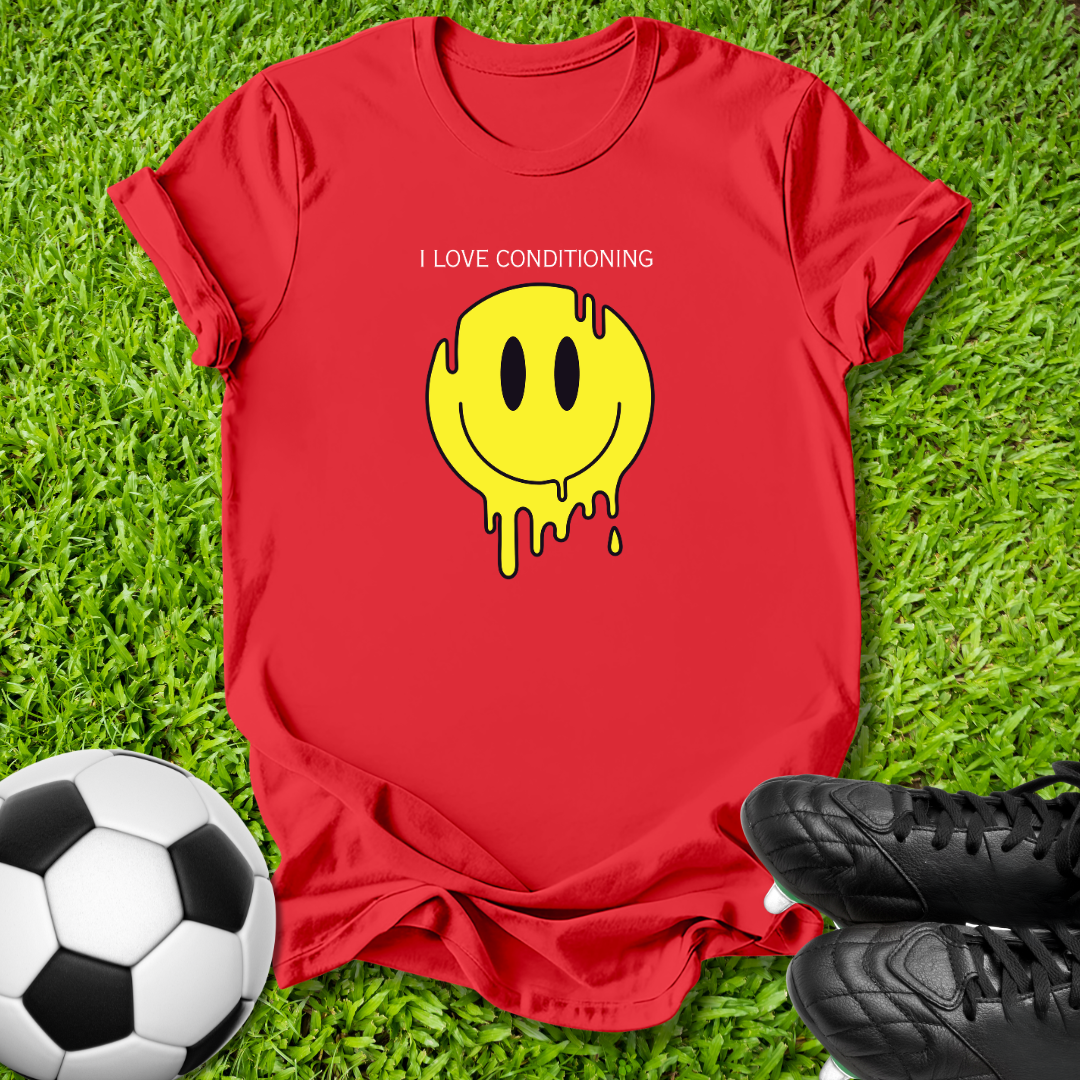 A red short-sleeve t-shirt laid flat on green grass, featuring a yellow dripping smiley face graphic and the text 'I LOVE CONDITIONING'. A soccer ball is visible on the left and black cleats are on the right, creating a casual outdoor flat-lay scene.