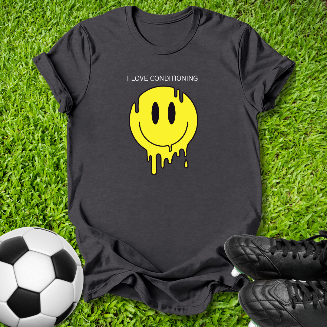 A black short-sleeve t-shirt laid flat on green grass, featuring a yellow dripping smiley face graphic with the text 'I LOVE CONDITIONING' above it. A soccer ball is visible on the left and black cleats are on the right.