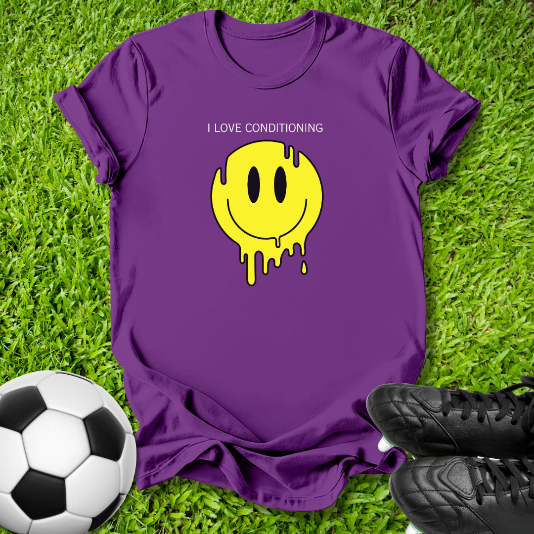 A purple short-sleeve t-shirt laid flat on green grass, featuring a yellow dripping smiley face graphic with the text 'I LOVE CONDITIONING' above it. A soccer ball is visible on the left and black cleats are on the right.