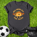 A black graphic t-shirt laid flat on green grass, featuring a cartoon taco character lifting dumbbells with the text 'WILL WORK OUT FOR TACOS'. A soccer ball is on the left and black cleats are on the right.