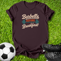 A dark brown short-sleeve t-shirt with a retro graphic reading 'Barbells BEFORE Breakfast' in beige and orange, styled on green grass with a soccer ball and black cleats.