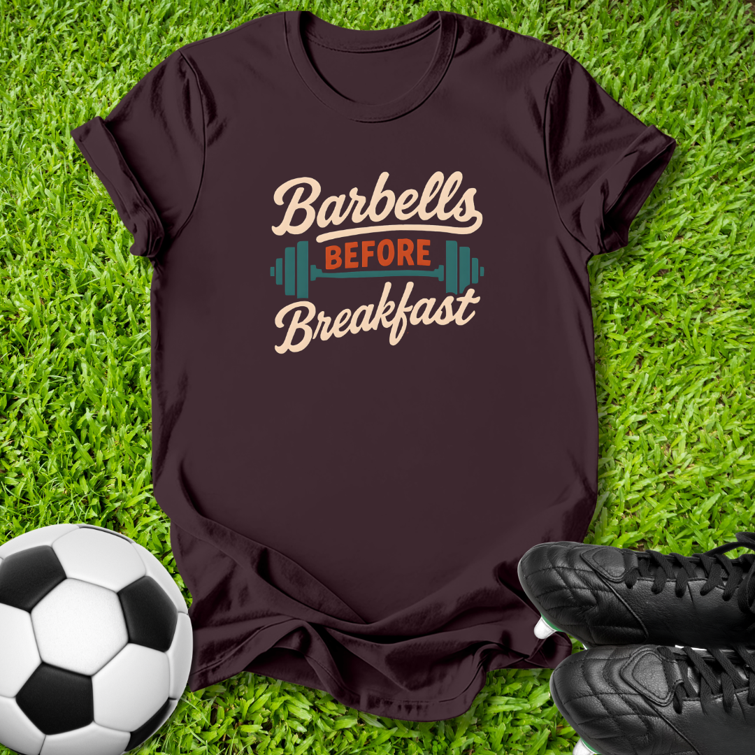 A dark brown short-sleeve t-shirt with a retro graphic reading 'Barbells BEFORE Breakfast' in beige and orange, styled on green grass with a soccer ball and black cleats.
