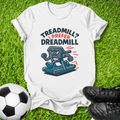 A white graphic t-shirt laid flat on green grass, featuring a cartoon treadmill character and the text 'TREADMILL? I PREFER DREADMILL'. A soccer ball is on the left and black cleats are on the right.