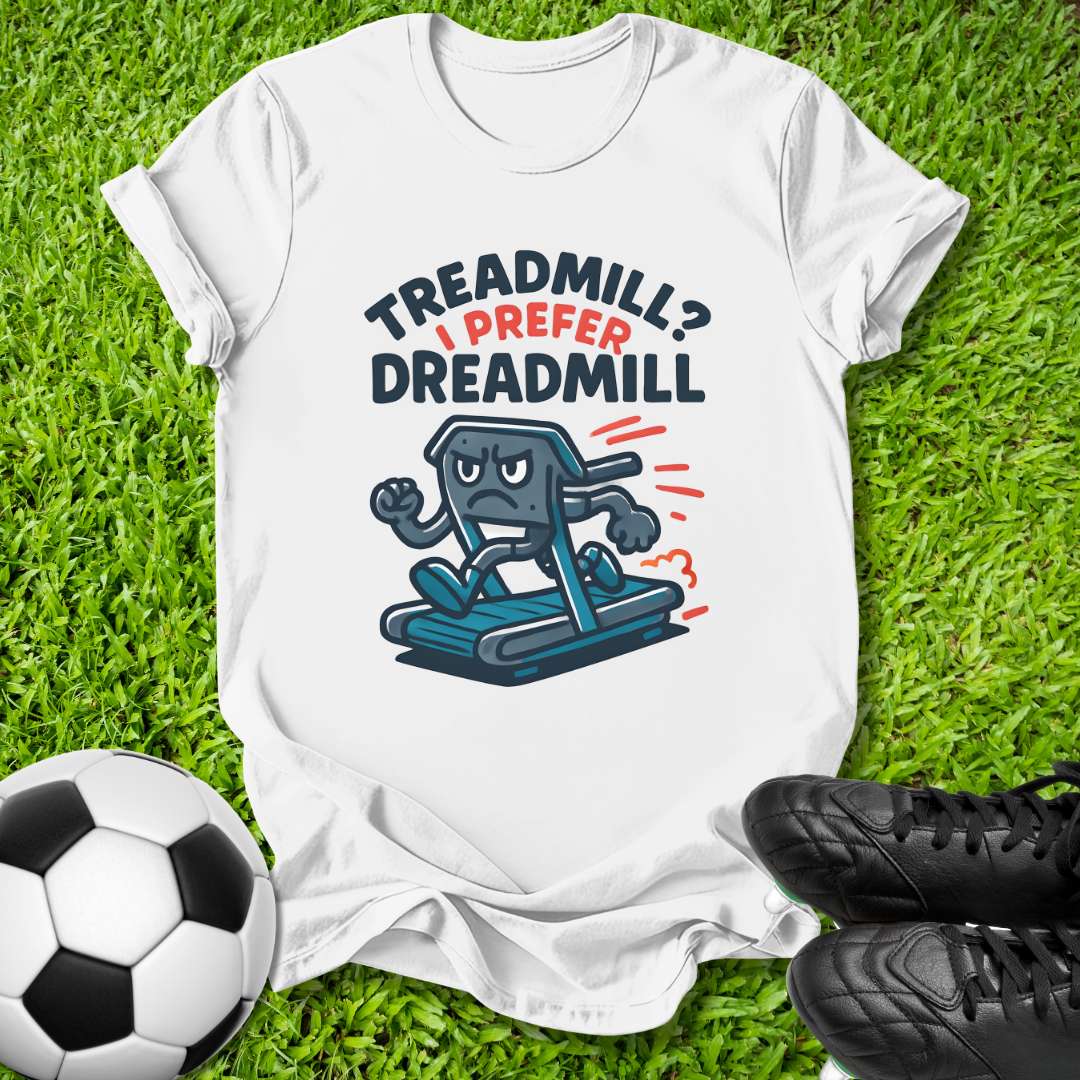 A white graphic t-shirt laid flat on green grass, featuring a cartoon treadmill character and the text 'TREADMILL? I PREFER DREADMILL'. A soccer ball is on the left and black cleats are on the right.