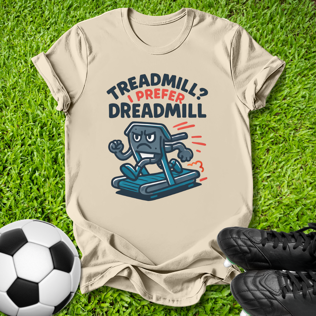A beige graphic t-shirt laid flat on green grass, featuring the text 'TREADMILL? I PREFER DREADMILL' above a cartoon treadmill character. A soccer ball is on the left and black cleats are on the right.