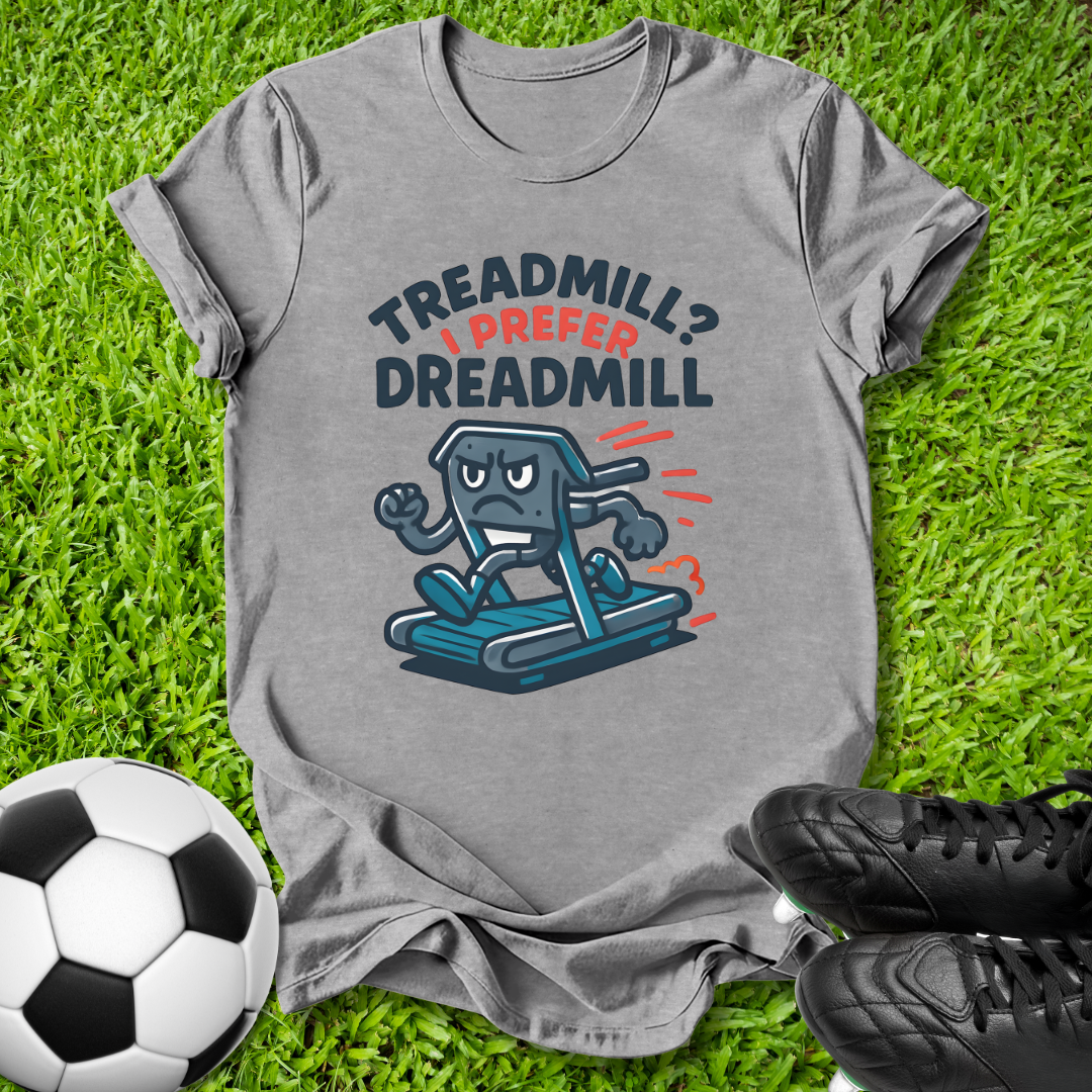 A heather gray graphic t-shirt laid flat on green grass, featuring a cartoon treadmill character and the text 'TREADMILL? I PREFER DREADMILL'. A soccer ball and black cleats are placed around it as props.