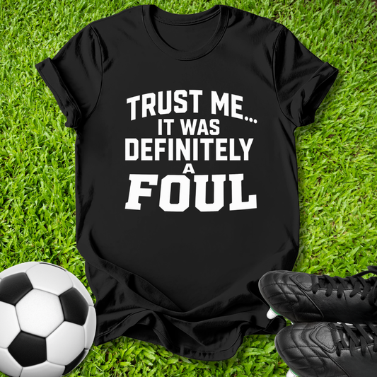A black short-sleeve graphic T-shirt laid flat on green grass, with bold white text reading 'TRUST ME.